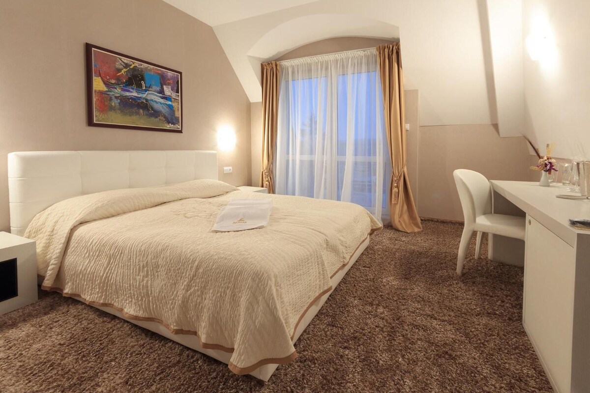 A hotel room with a bed, a desk, a chair, a painting on the wall, and a window with curtains.