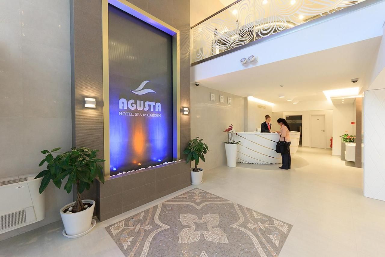 Zona de recepción de un hotel con diseño moderno, que incluye un mostrador de recepción, plantas en macetas y una pared decorativa con un logotipo.