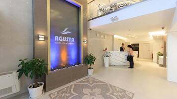 Zona de recepción de un hotel con diseño moderno, que incluye un mostrador de recepción, plantas en macetas y una pared decorativa con un logotipo.