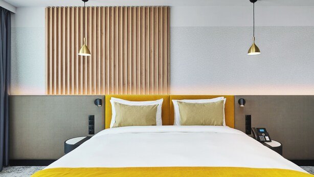 Quarto de hotel moderno com uma cama grande, duas mesas de cabeceira, um telefone fixado na parede e duas luminárias pendentes.
