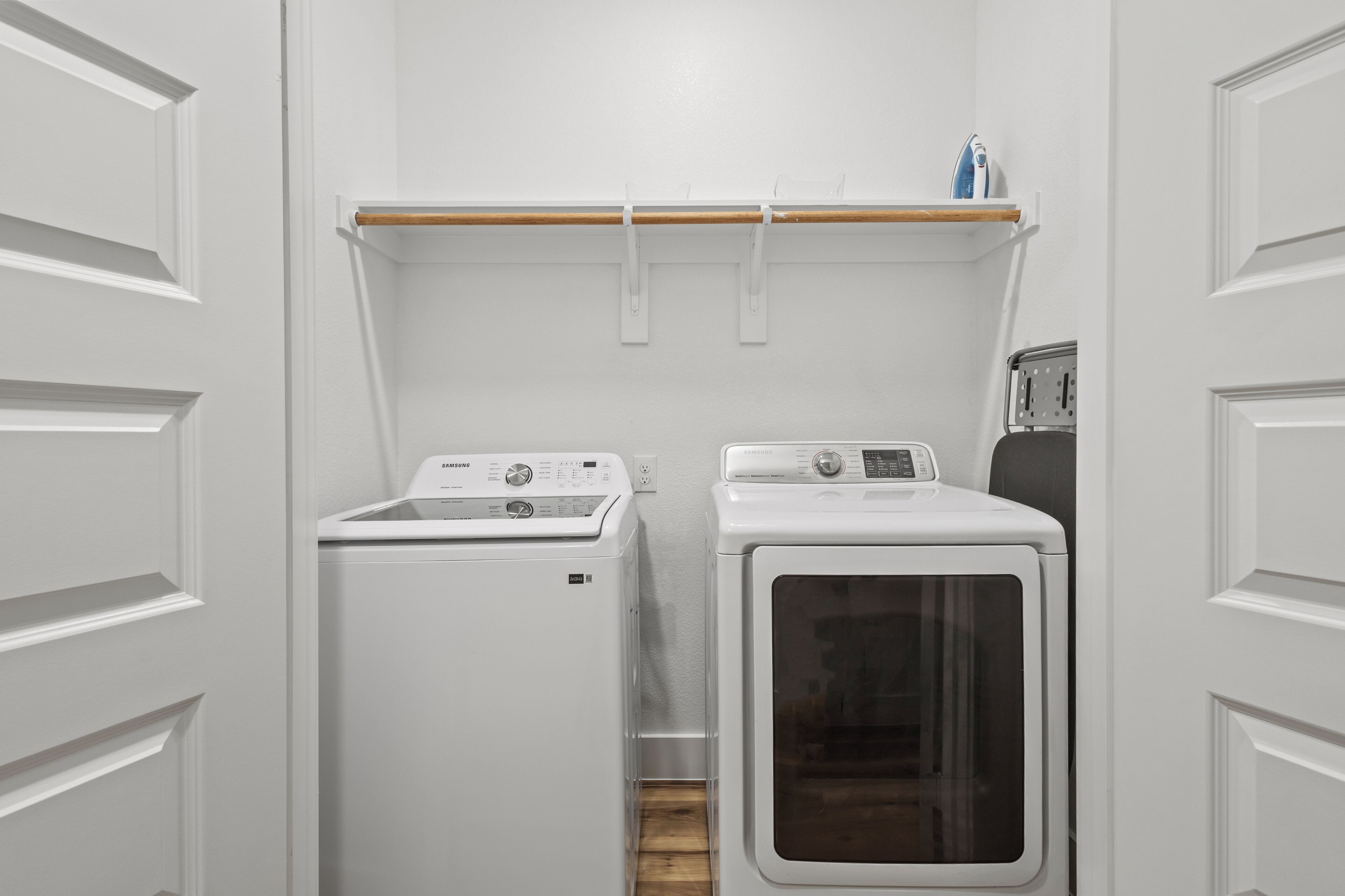 A laundry area with a front-loading washer and dryer, a wooden shelf above, and a wall-mounted ironing board.