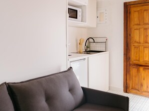 Een kleine keuken met een magnetron, spoelbak en vaatwasser, een houten deur en een grijze bank.
