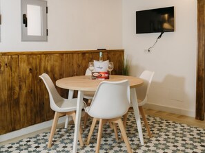 Un coin repas avec une table ronde, des chaises blanches, une bouteille de vin et un téléviseur fixé au mur.