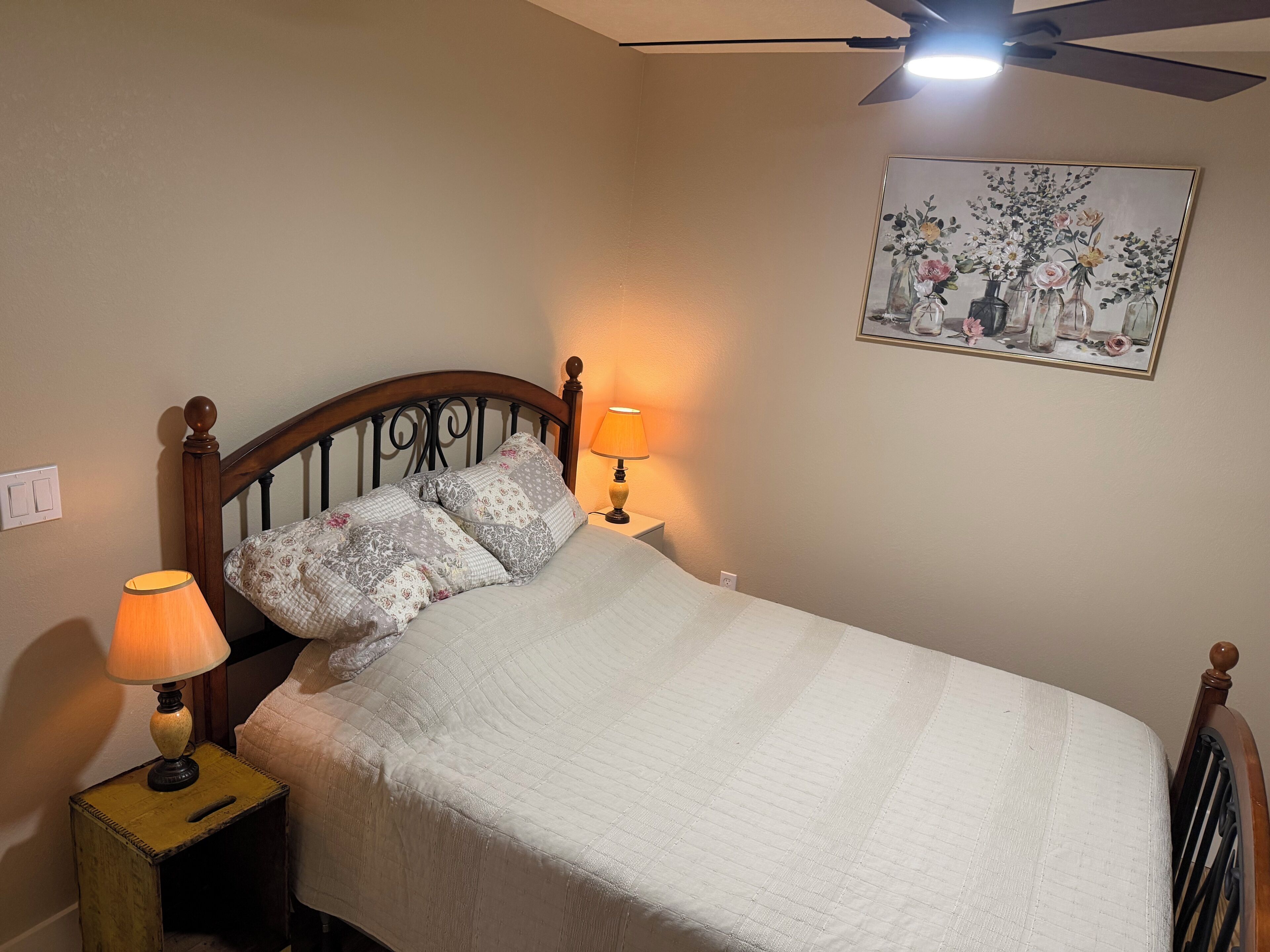 A neatly made bed with a white comforter, two bedside lamps, a ceiling fan, and a floral painting on the wall.