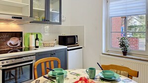 Una cocina con estufa, microondas y armarios, una mesa de comedor con un mantel individual, y una ventana con vista a un edificio de ladrillo.