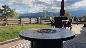 A circular outdoor fire pit with flames, surrounded by a patio area with chairs and an umbrella, set against a backdrop of mountains and a cloudy sky. - Mountain Views, Air Force Academy, Hot Tub, Pet Friendly, Hiking, Trails, Dining (Colorado Springs)