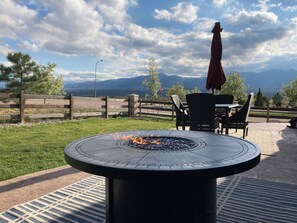 A circular outdoor fire pit with flames, surrounded by a patio area with chairs and an umbrella, set against a backdrop of mountains and a cloudy sky. - Mountain Views, Air Force Academy, Hot Tub, Pet Friendly, Hiking, Trails, Dining (Colorado Springs)