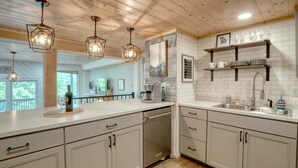 A modern kitchen with white cabinetry, a built-in dishwasher, and a coffee maker. There are pendant lights hanging from the ceiling and a window providing natural light. - Summit View - Mountain Top View & Gas Fireplace (Hiawassee)