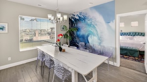 Un coin repas avec une table blanche, des chaises transparentes, un lustre et une grande fresque murale représentant une vague.