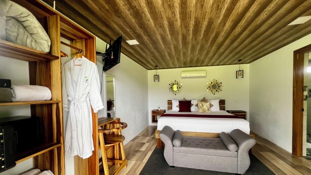 Quarto com teto de madeira, cama com colcha vermelha e dourada, sofá cinza, prateleira de madeira com toalhas e uma televisão na parede.