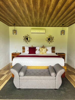 A bedroom with a wooden ceiling, two wall-mounted air conditioners, a bed with white bedding and decorative pillows, bedside tables, and a grey upholstered bench. - Romantic Arenal Escape : Jacuzzi + King Bed : Villa Luna 3 (La Fortuna)