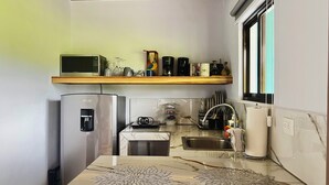 Cozinha compacta com micro-ondas, frigorífico, máquina de café e janela com persianas.