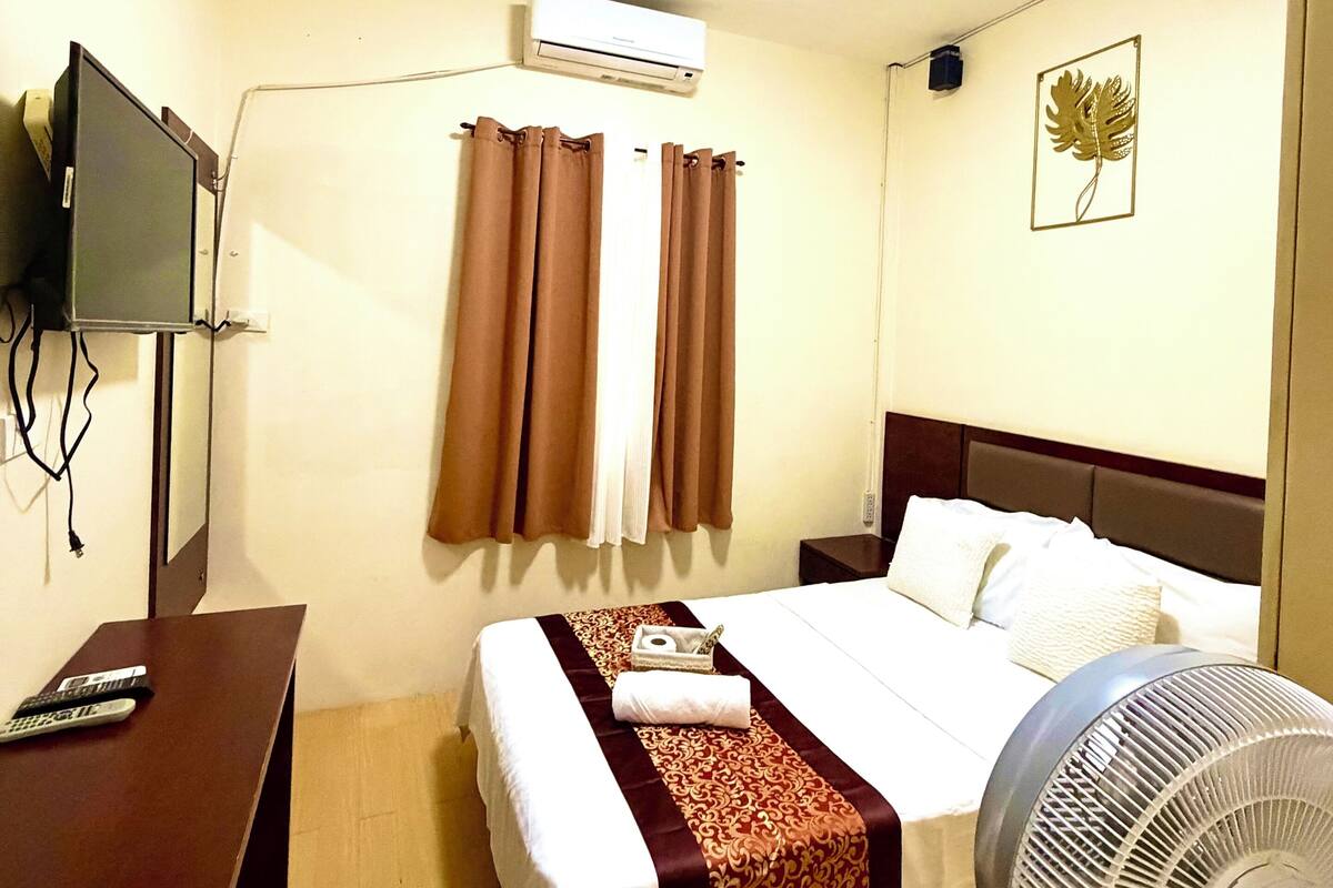 Habitación de hotel con cama, escritorio, televisor, aire acondicionado, ventilador y una obra de arte colgada en la pared.