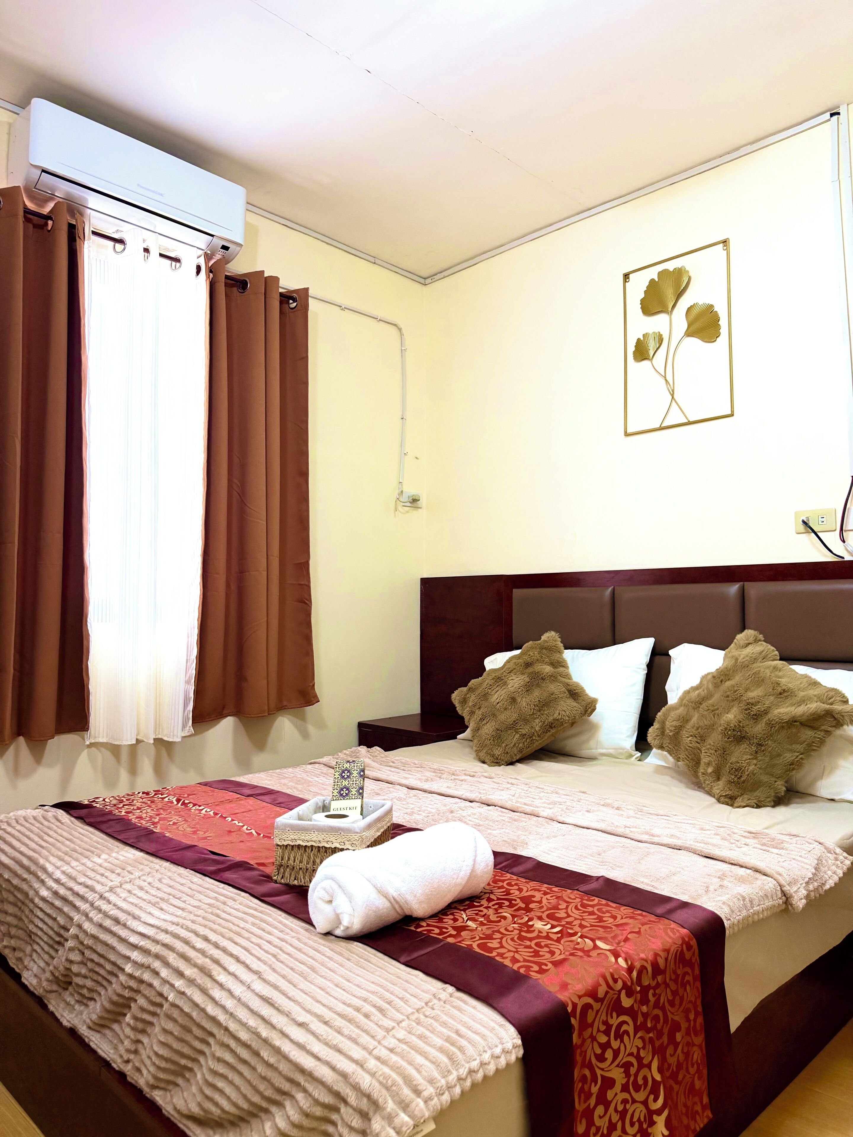 Habitación de hotel con cama, cortinas marrones, aire acondicionado de pared, mesita de noche y un adorno en la pared.