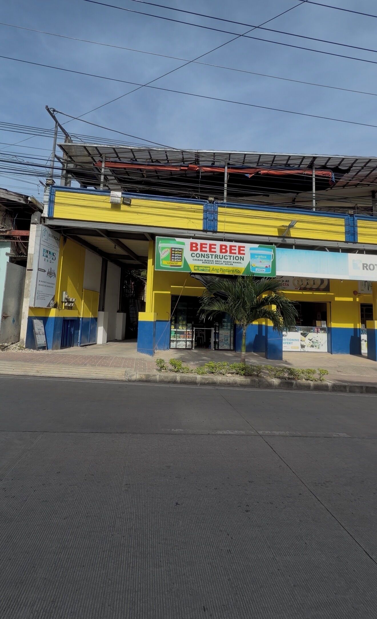 A commercial building with a yellow and blue facade, a sign for 'BEE BEE CONSTRUCTION,' and a palm tree in front.