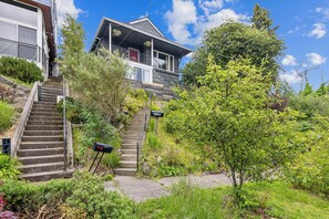 A residential house with a staircase leading to a porch, surrounded by lush greenery and a clear blue sky. - Spring Haven Lakeside Casita | Quick Trip Downtown (Seattle)