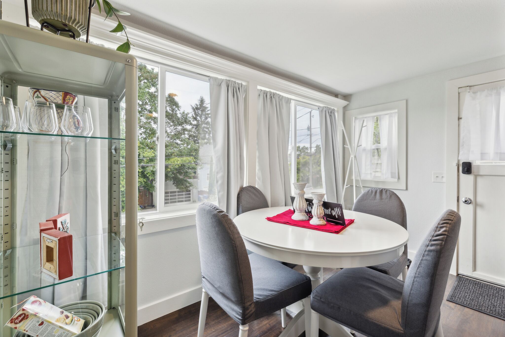 A dining area with a round table, grey upholstered chairs, and a glass display unit with various items.