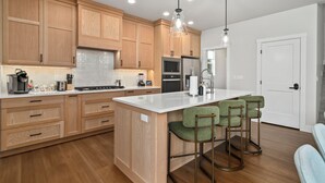 A modern kitchen with wooden cabinets, a central island, bar stools, and pendant lighting. - Pro Golf Simulator and Hot Tub, New Luxury 4 BR, Boise Home Sleeps 12 (Boise)