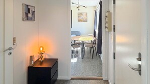 Un passadís que condueix a una zona de menjador amb una taula rodona i cadires, un lluminari fixat a la paret i una porta amb mànec.