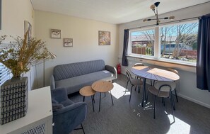 A modern living room with a grey sofa, round coffee table, and dining table with chairs. There are framed artworks on the wall and a large window with curtains.