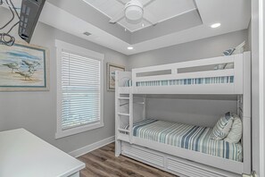 Quarto com beliche, ventilador de teto, janela com persianas e um quadro na parede.