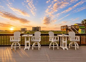 A deck with four Adirondack chairs and small tables, overlooking a sunset and buildings. - LCV33 Shared Pool, Private Hot Tub,Propane Grill (Port Aransas)