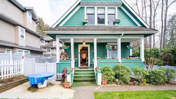 A two-story house with a green exterior, a front porch with steps, and a small front yard with a bench and potted plants.