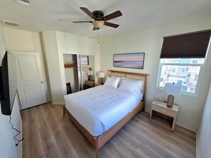 Ein Hotelzimmer mit einem Bett, einem Deckenventilator, einem Nachttisch, einer Lampe, einem Fenster mit Jalousien und einem Bild an der Wand.