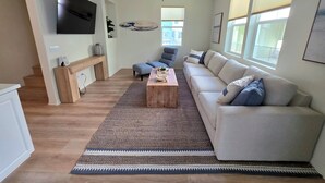 Uma sala de estar moderna com uma televisão de tela plana, um ventilador de teto, um sofá, uma mesa de centro e uma grande janela com persianas.