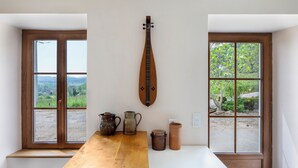 Ein Zimmer mit einem Holztisch, einem an der Wand befestigten Saiteninstrument und zwei Fenstern mit Blick auf Grünflächen.