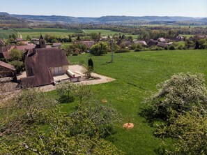 Paisaje rural con una casa grande que cuenta con una torre destacada, rodeada de campos verdes exuberantes y colinas distantes.