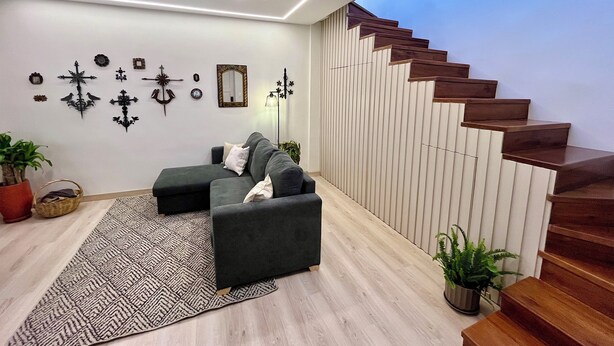 Ein modernes Wohnzimmer mit grauem Sofa, einem gemusterten Teppich und Wanddekoration. Eine Holztreppe führt zu einer oberen Ebene.
