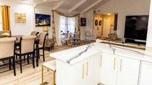 Eine moderne Küche mit weißen Schränken und goldenen Griffen, ein Essbereich mit Tisch und Stühlen sowie ein Wohnzimmer mit Fernseher und Sitzgelegenheiten.