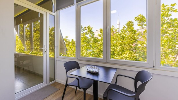 Una habitación con una mesa y sillas, ventanas amplias que dan a vistas de árboles y un edificio.