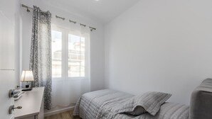 A bedroom with a bed, a bedside table, a lamp, a window with curtains, and a view of buildings outside. - Casa Tirana Cozy 2BR Apartment Near Main Square (Tirana)