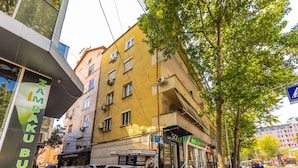 A street view with a yellow building, a sign for 'ZAMBUKO UTI,' and a pedestrian crossing. - Casa Tirana Cozy 2BR Apartment Near Main Square (Tirana)