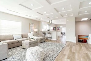 Ein modernes Wohnzimmer mit einer Couch, einem Hocker und einem gemusterten Teppich. Im Hintergrund befindet sich eine Küchenzeile mit Barhocker und einem Esstisch.