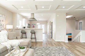 Ein modernes Wohnzimmer mit Sofa, Couchtisch und Teppich. Im Hintergrund ist eine Küchenzeile mit Bar und Hockern zu sehen.