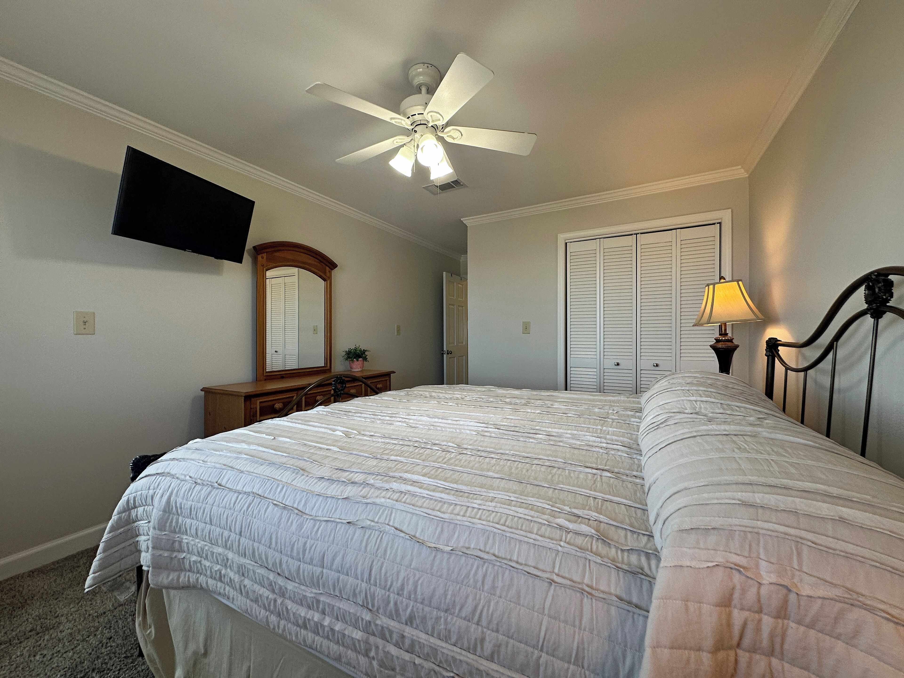 A bedroom with a bed, a ceiling fan, a TV mounted on the wall, a dresser, a mirror, a lamp, and a closet.