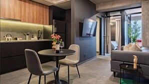 Ein modernes Wohnzimmer mit einer Couch, einem Esstisch mit Stühlen, einer Küchenzeile inklusive Kühlschrank und einem an der Wand montierten Fernseher.