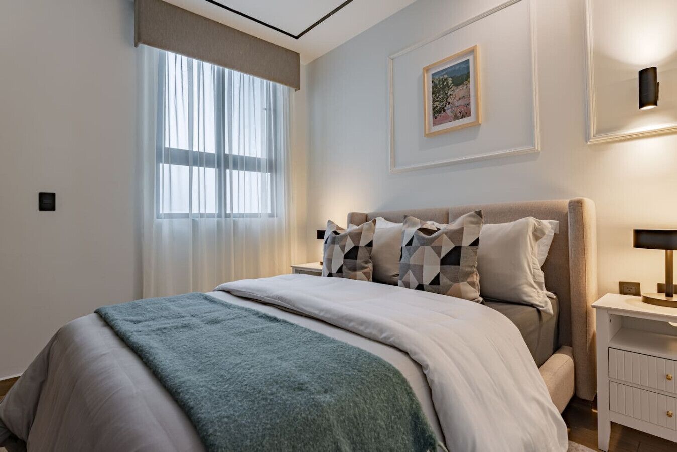 A bedroom with a bed, a nightstand, a lamp, a window with curtains, and a framed picture on the wall.