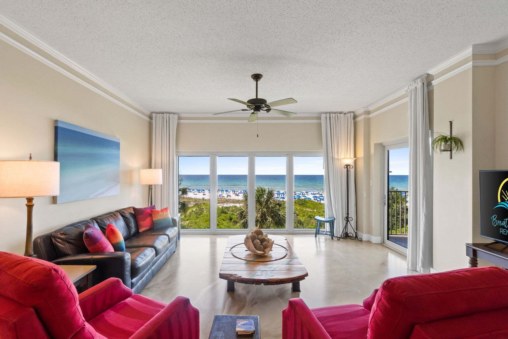 A living room with a view of the beach, a ceiling fan, and a television.