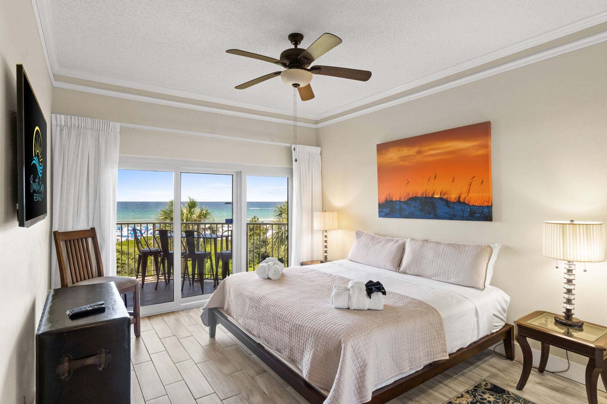 Une chambre avec un lit, un ventilateur de plafond, une télévision, un balcon avec vue sur l’océan et un tableau au mur.