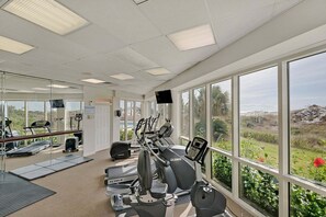 Ein Fitnessraum mit Cardiogeräten, einem Fernseher und großen Fenstern mit Blick auf Grünflächen.
