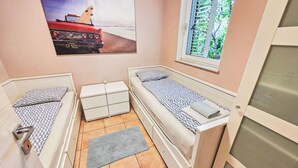 Una stanza compatta con due letti singoli, un tavolino, una finestra con persiane e un quadro a tema spiaggia.