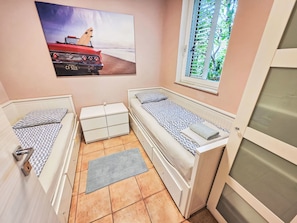 Ein gemütlicher Raum mit zwei Einzelbetten, einem kleinen Tisch, einem Fenster mit Jalousien und einem Strandmotiv an der Wand.