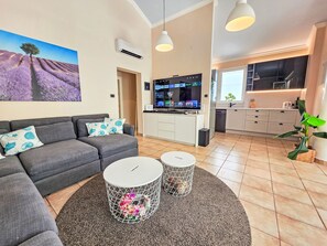 Ein modernes Wohnzimmer mit einem grauen Sofa, einem Flachbildfernseher und einer Küchenzeile mit Fenster.