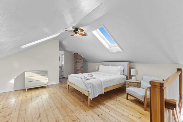 A bedroom with a sloped ceiling, a skylight, a bed, a chair, and a small table.