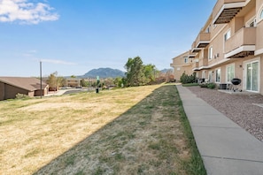 A residential area with a grassy lawn, a paved walkway, and a row of beige apartment buildings with balconies. - 1BR Cozy Studio, Patio, Grill, Near Garden of Gods (Colorado Springs)