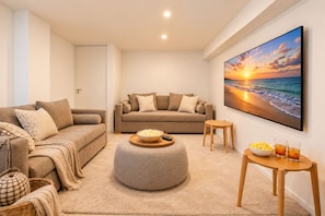Ein modernes Wohnzimmer mit einem beigefarbenen Sofa, einem runden Hocker, einem kleinen Holztisch mit Snacks und einem großen Gemälde, das einen Sonnenuntergang über dem Meer zeigt.